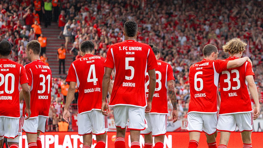 Die Spieler des 1. FC Union Berlin in roten Trikots stehen auf dem Spielfeld vor jubelnden Fans im Hintergrund.