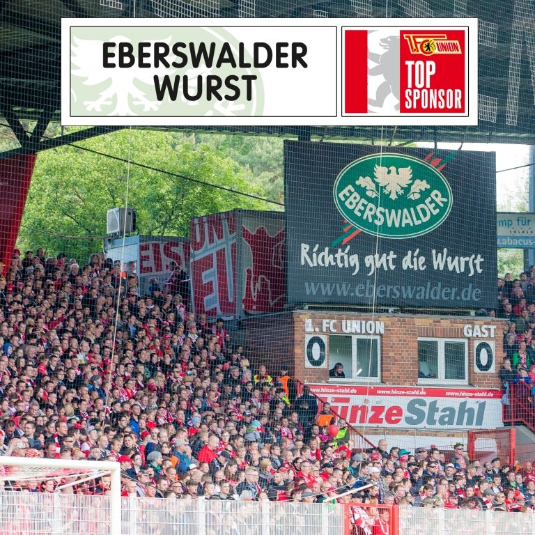 Voller Fußballstadion mit Zuschauern, großem Eberswalder-Wurst-Schild und Sponsor-Logos oben.