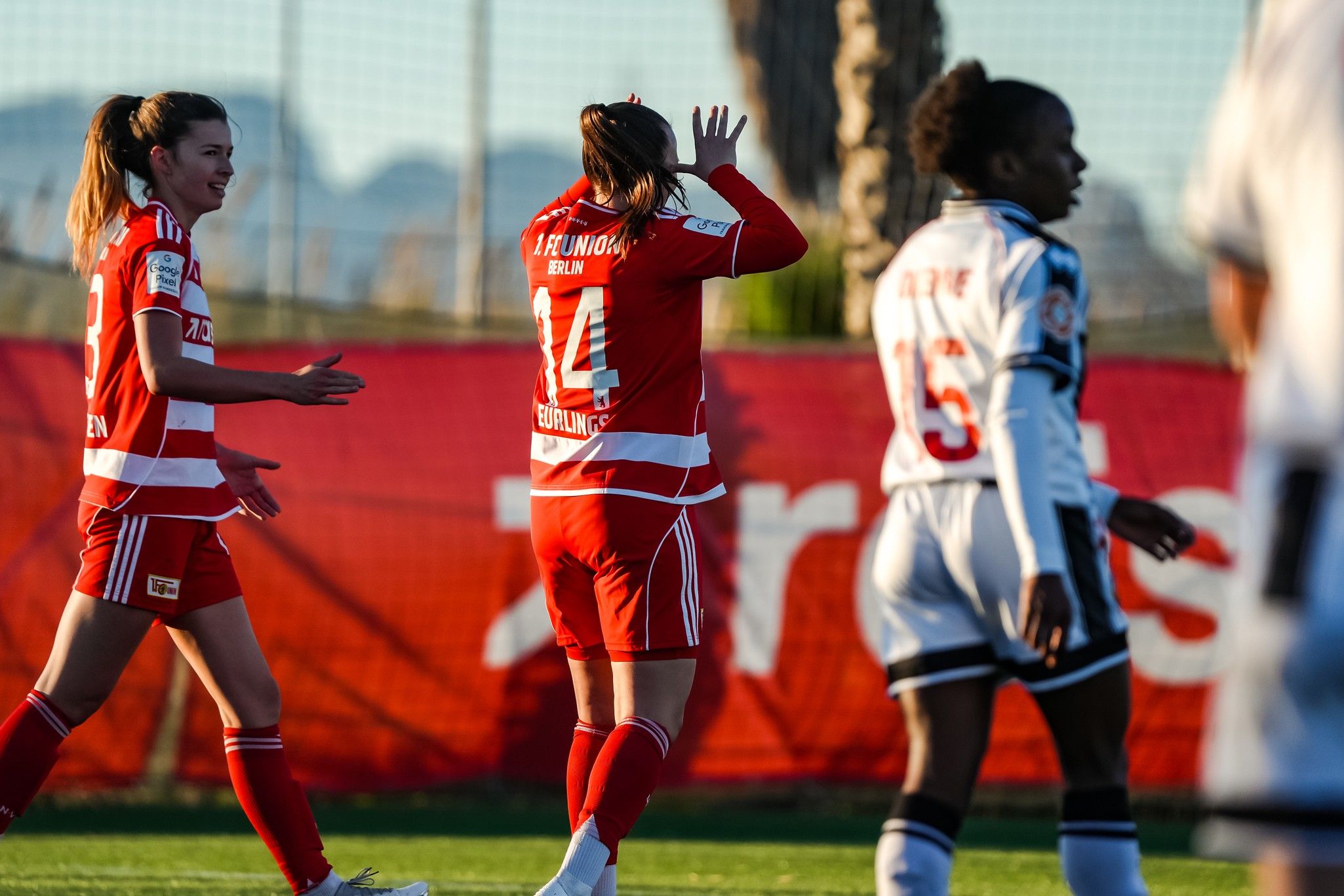 Zwei Fußballspielerinnen in roten und weißen Trikots feiern, während eine dritte Spielerin in dunklem Trikot am Spiel teilnimmt.