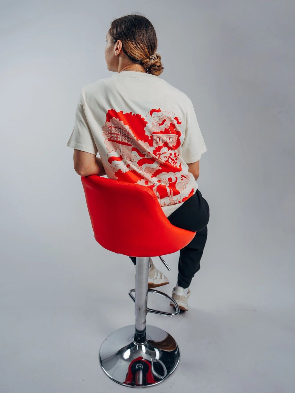 Eine Person sitzt auf einem roten Barstuhl und trägt ein T-Shirt mit einem bunten, grafischen Muster auf dem Rücken.