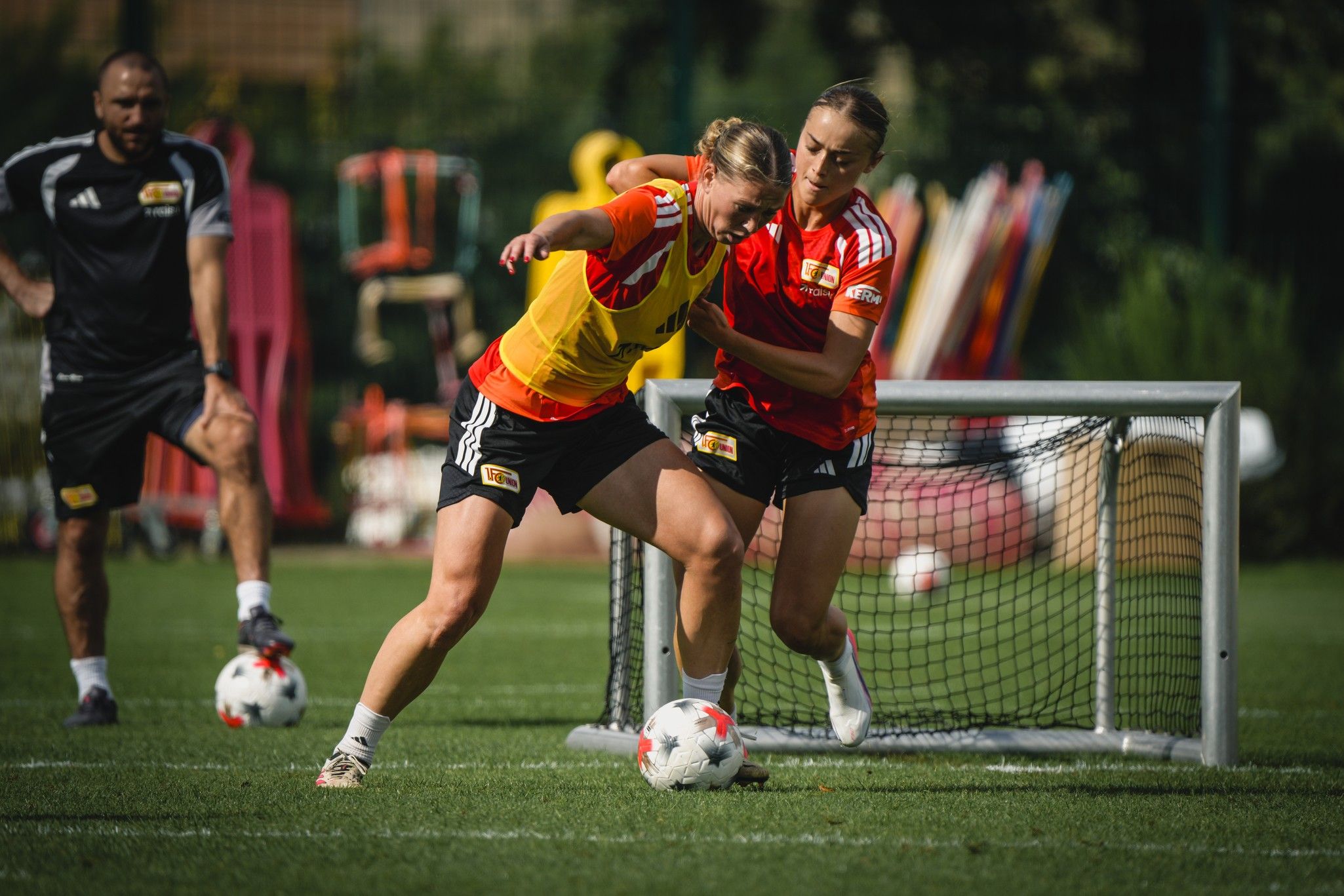 Zwei Fußballspielerinnen in Trainingssituation, eine nimmt die andere in den Schwitzkasten, mit einem Tor im Hintergrund.