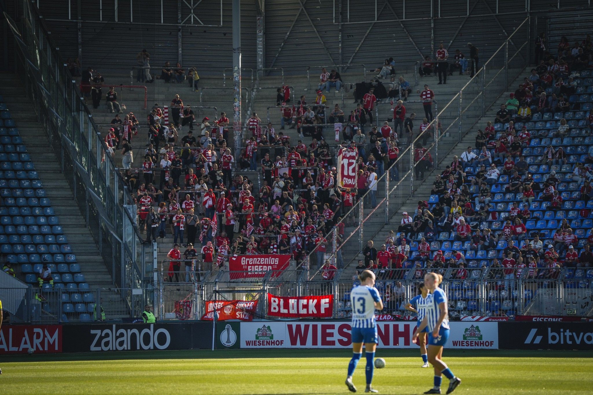 Zwei Fußballspieler stehen auf dem Rasen, während fanatische Zuschauer mit Bannern auf den Tribünen stehen.