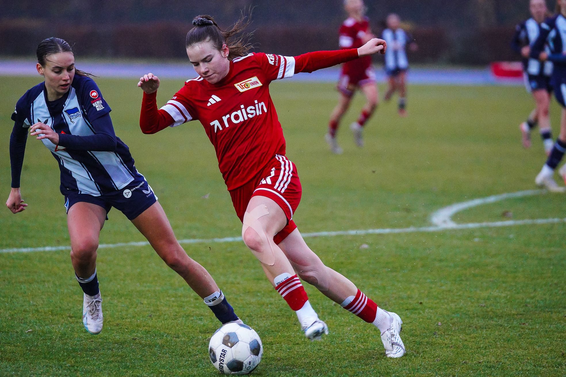 Zwei Frauen im Fußballspiel, eine in roter und die andere in blauer Uniform, kämpfen um den Ball auf einem Rasenplatz.