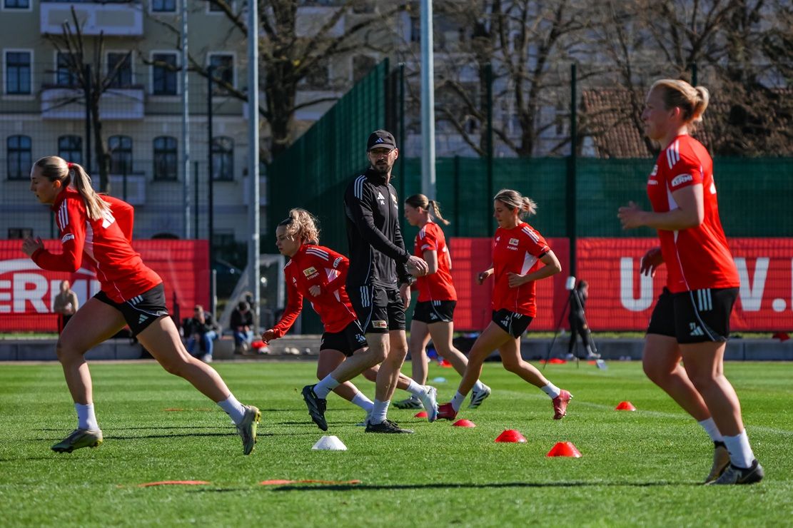 Ein Trainer beobachtet eine Gruppe von Frauen beim Fußballtraining auf einem Rasenfeld mit Markierungen und Anhängern im Hintergrund.