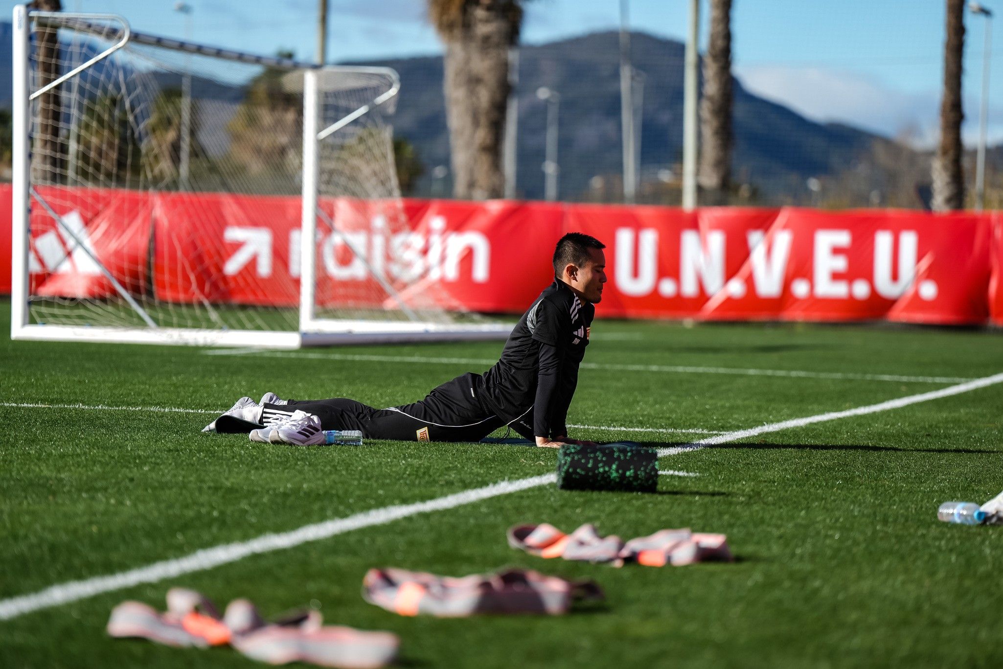 Ein Fußballspieler dehnt sich auf dem Trainingsplatz, umgeben von Sportequipment und einem Tor im Hintergrund.