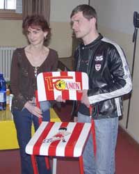 Zwei Personen halten einen Stuhl mit rot-weißen Streifen und einem Vereinslogo. Hintergrund unscharf mit Gelb- und Brauntönen.