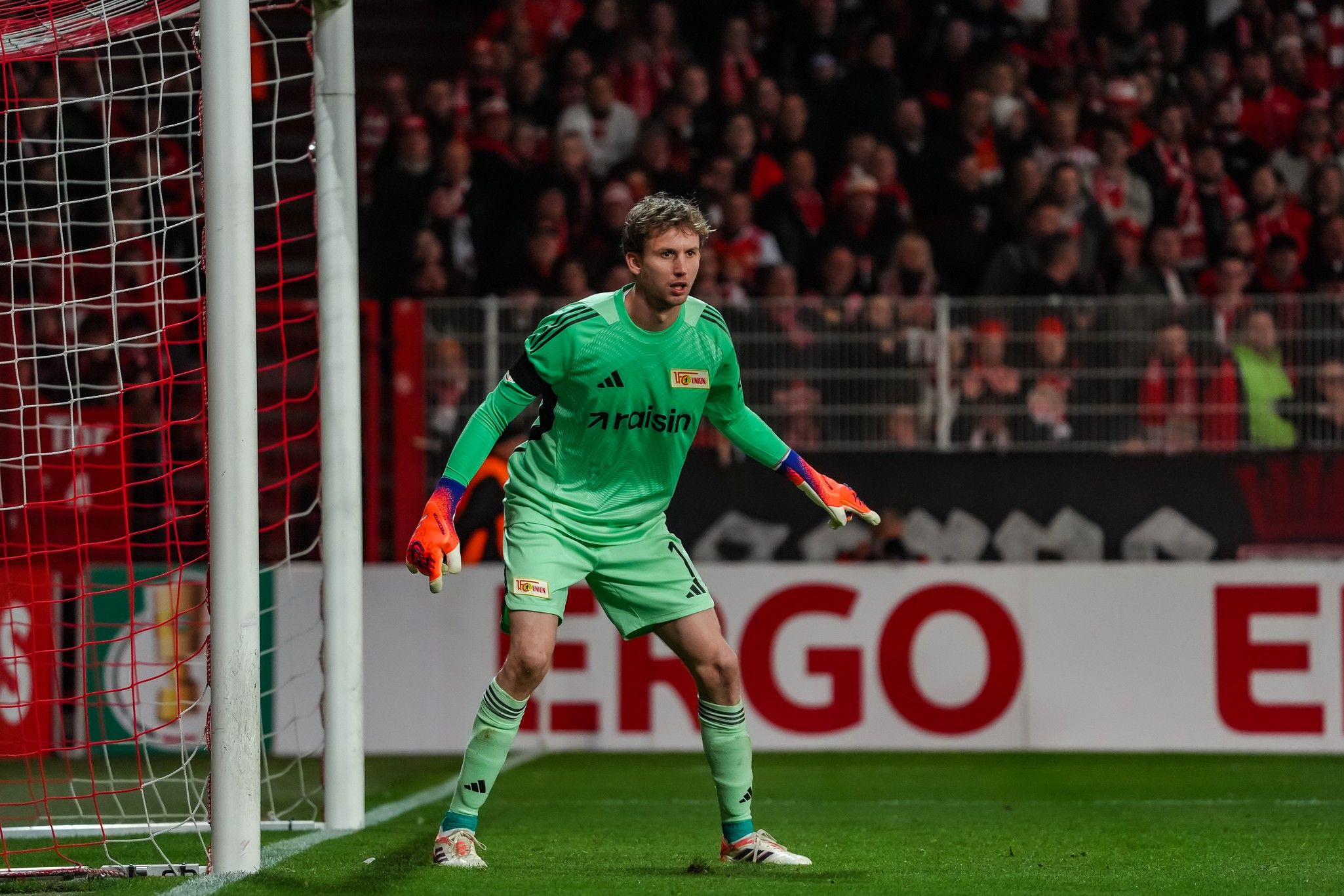 Ein Torwart in grüner Uniform steht im Tor, bereit, einen Schuss abzuwehren, während Zuschauer im Hintergrund zuschauen.