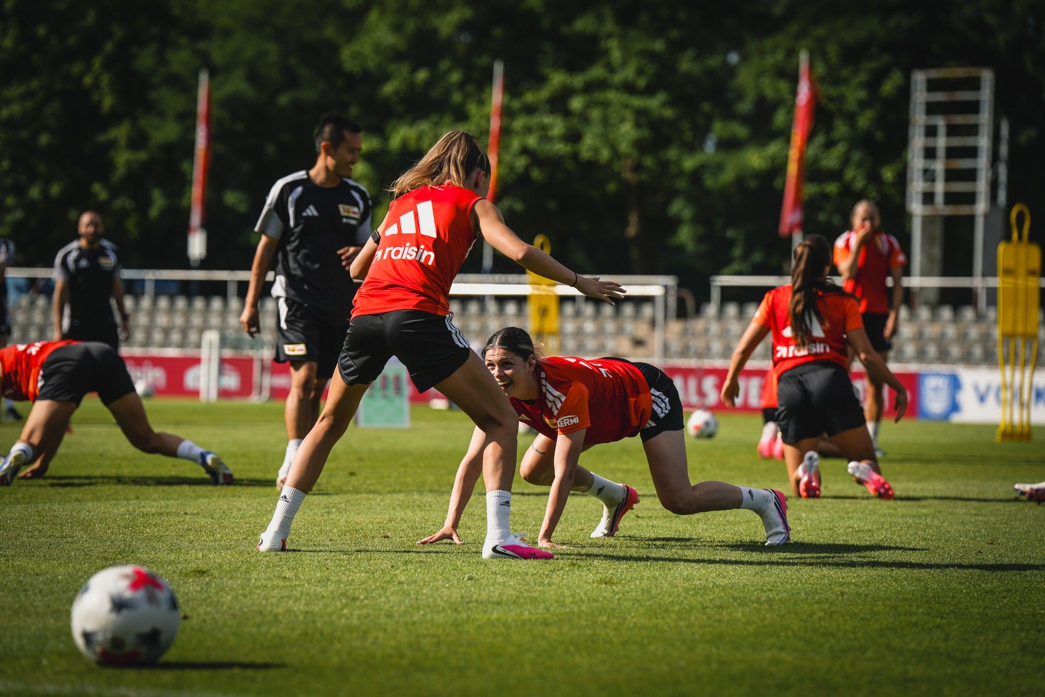 Zwei Spielerinnen trainieren auf einem Fußballplatz, während andere im Hintergrund sich ebenfalls aufwärmen.