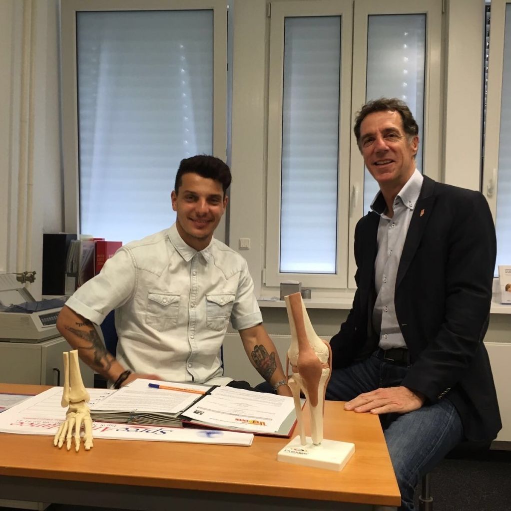 Zwei Männer sitzen an einem Tisch mit medizinischen Modellen eines Hand- und Kniegelenks. Sie lächeln in die Kamera.