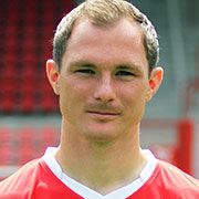 Ein lächelnder Mann in einem roten Fußballtrikot vor einer sportlichen Kulisse.