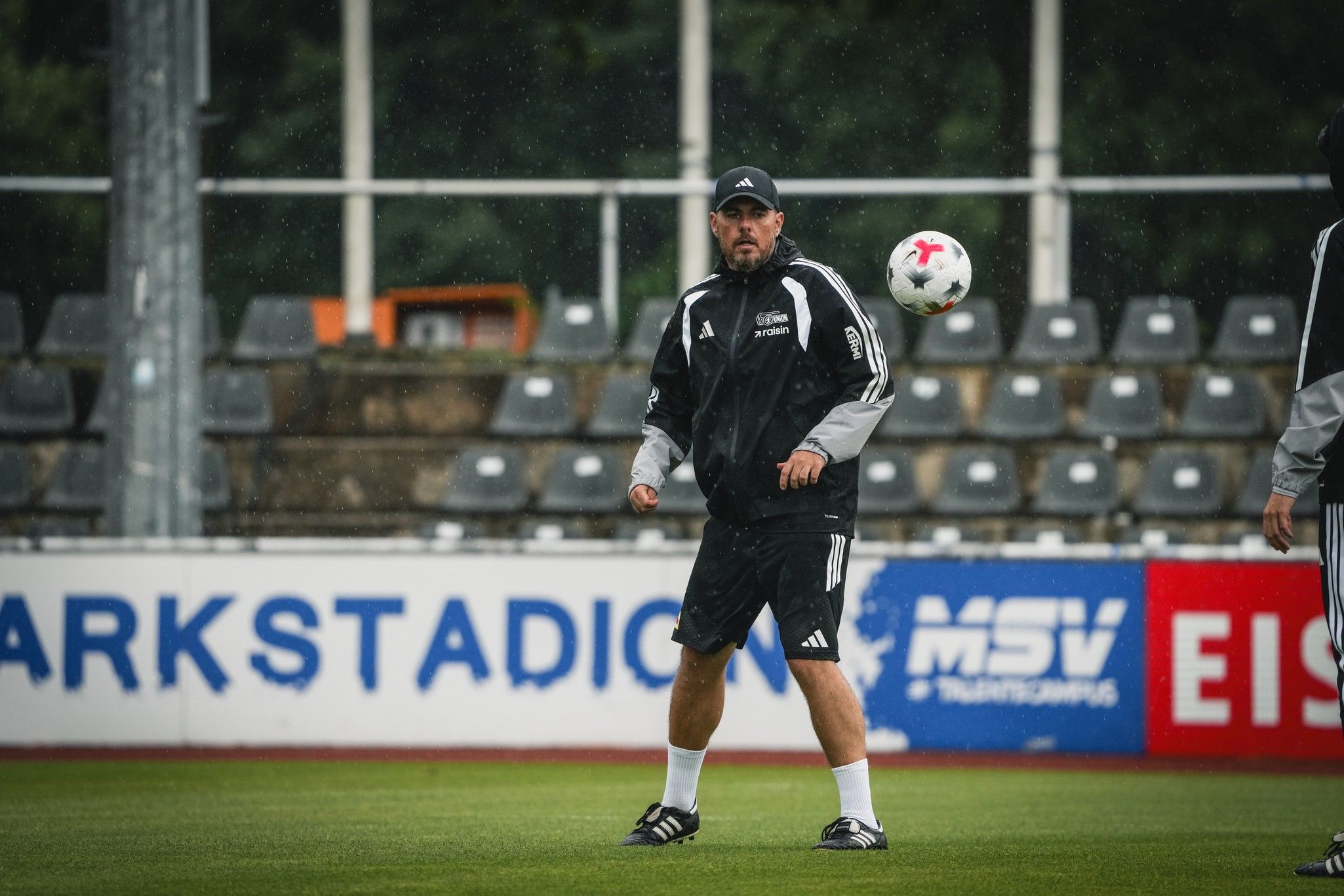 Ein Fußballtrainer in Trainingskleidung fängt einen Ball auf einem Platz unter regnerischem Wetter.