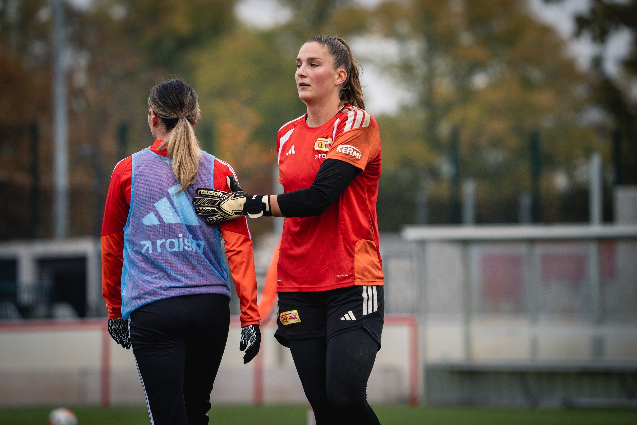 Zwei Fußballspielerinnen im Training, eine in einem roten Trikot mit Handschuhen, die andere mit einer blauen Weste.