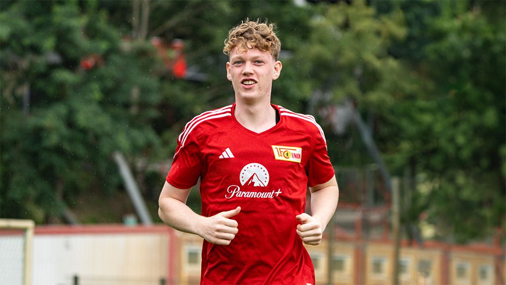 A young soccer player in a red jersey jogs smiling on a training field, surrounded by trees and a blurred background.