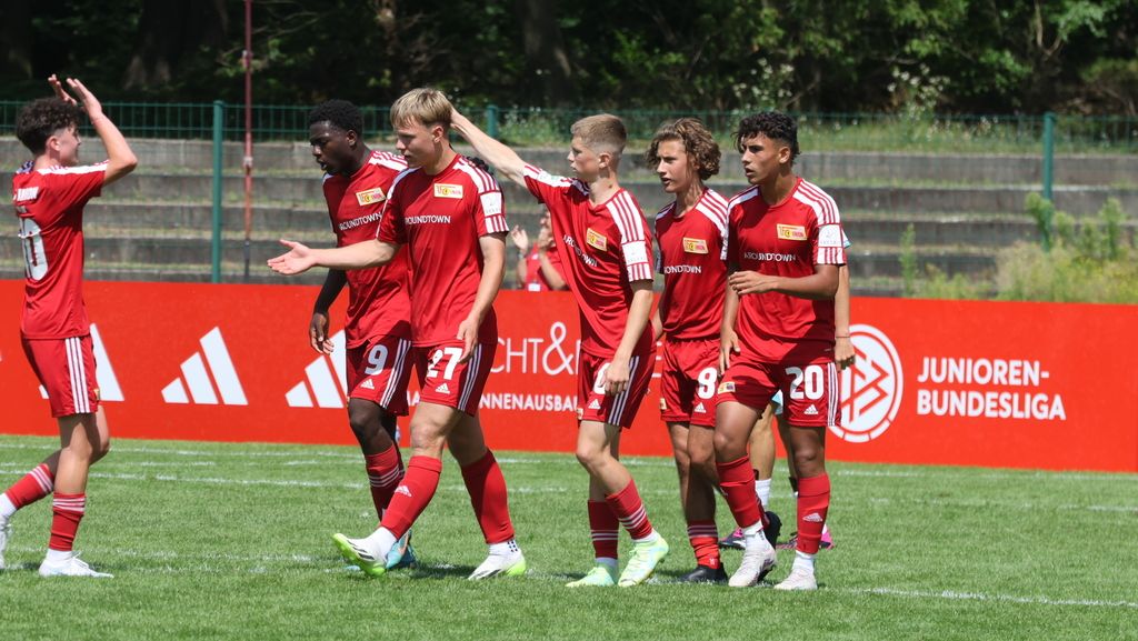 Sechs jubelnde Fußballspieler in roten Trikots feiern auf dem Spielfeld zusammen. Im Hintergrund ein Banner der Junioren-Bundesliga.