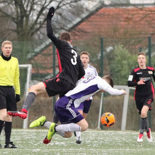 Ein Fußballspieler fällt nach einem Zweikampf auf das Spielfeld, während ein Schiedsrichter und weitere Spieler im Hintergrund stehen.