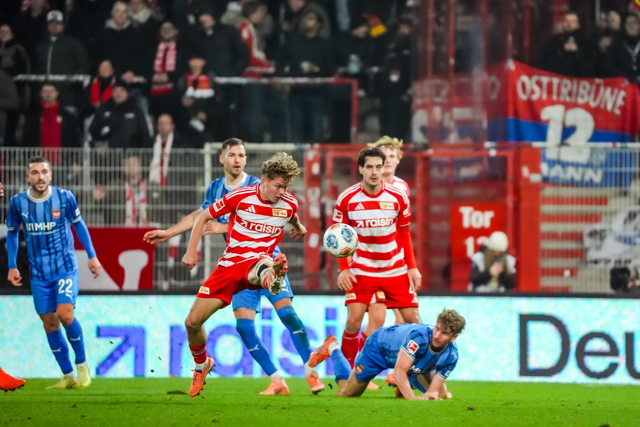 Fußballspiel mit Spielern in rot-weißen und blauen Trikots, ein Spieler fällt zu Boden, während andere in Aktion sind.