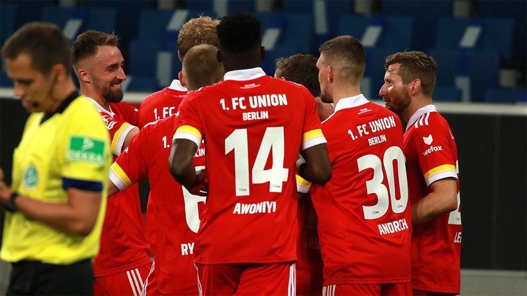 Spieler vom 1. FC Union Berlin freuen sich. Sie haben ein Tor geschossen. Sie tragen rote Trikots. Sie sind auf dem Spielfeld.