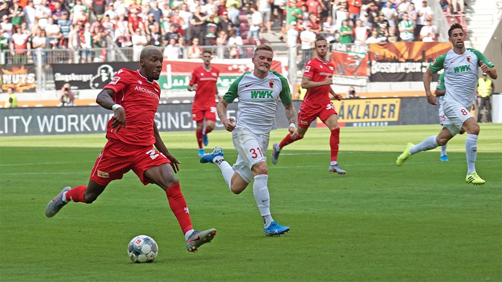 Game scene from a football match where a player in a red jersey dribbles the ball while an opponent in white chases him.