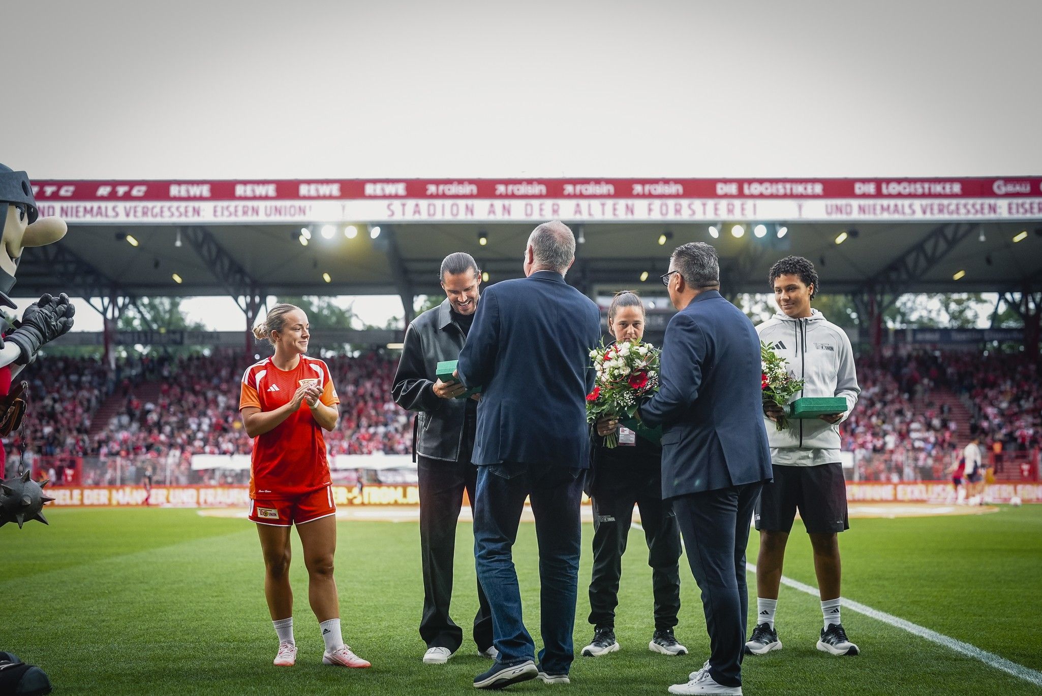 Ein Fußballspiel mit einer Spielerin in rotem Trikot, die einer ceremonielle Ehrung beiwohnt, umgeben von Personen mit Blumen.