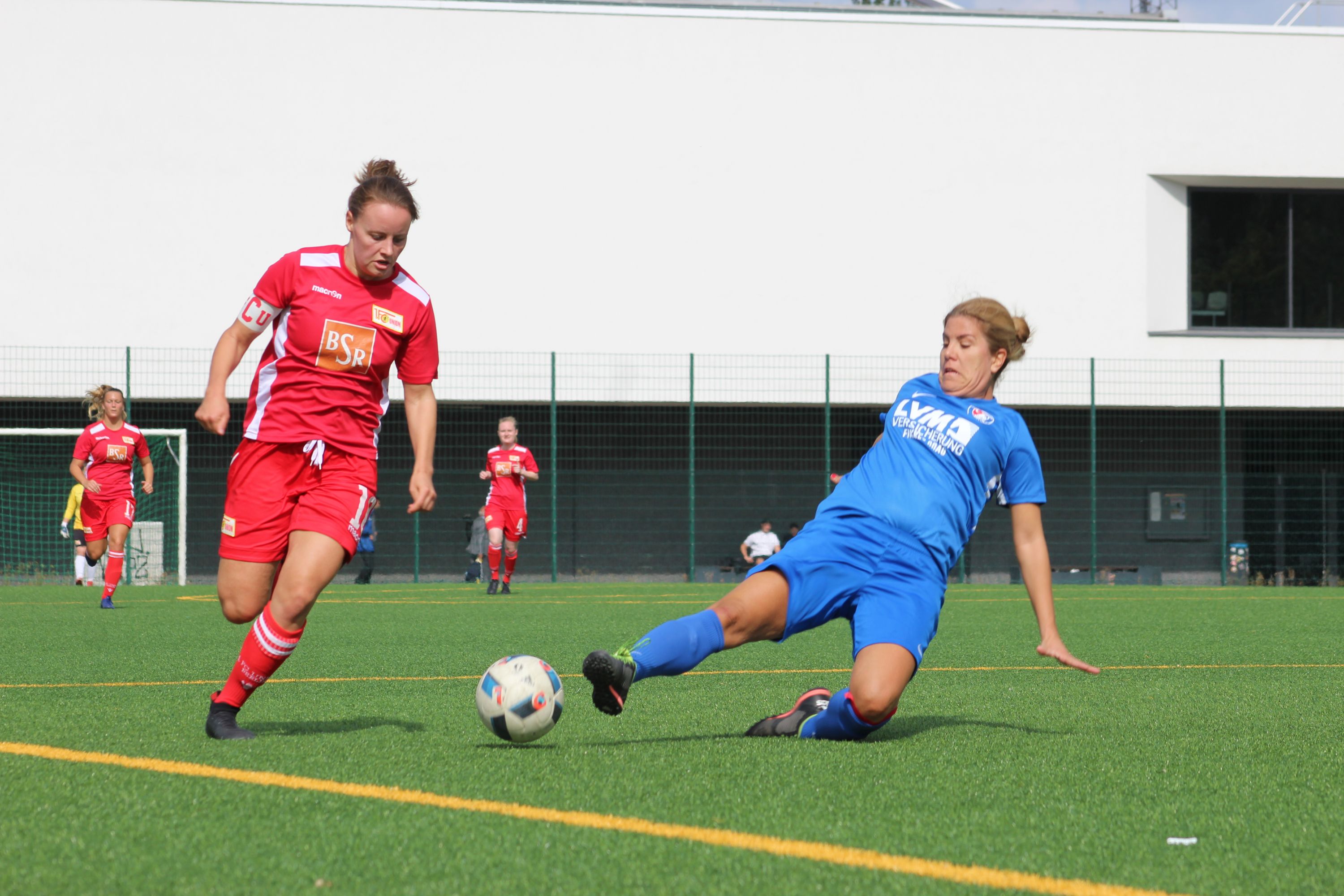 Zwei Fußballspielerinnen auf dem Platz; eine in rotem Trikot dribbelt, während die andere in Blau rutschend versucht, den Ball zu erreichen.