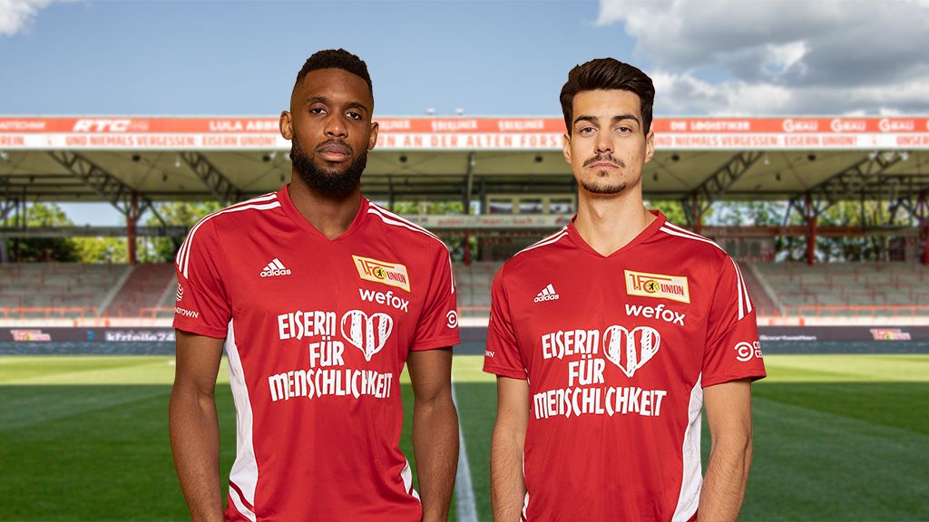 Two football players in red jerseys are standing on a stadium field, with stands and trees visible in the background.