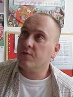 Ein Mann mit kurzem, glattem Haar und Ohrstecker schaut nach oben, vor einer Wand mit Bildern und Notizen.