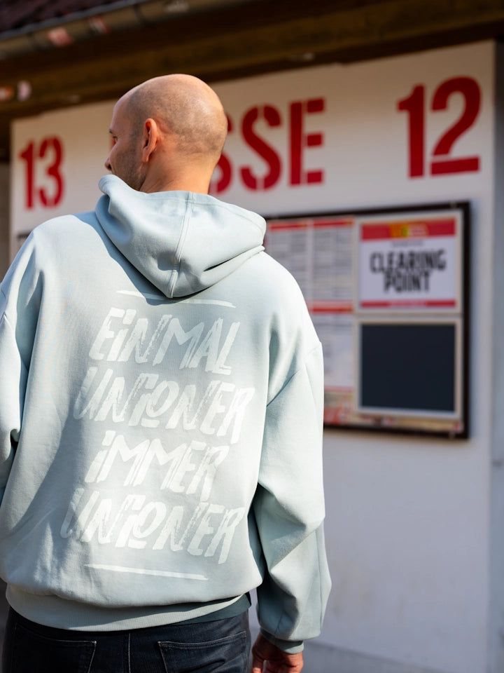 Mann mit Glatze trägt grauen Hoodie mit Schriftzug, steht vor einer Wand mit Plan und roten Zahlen.