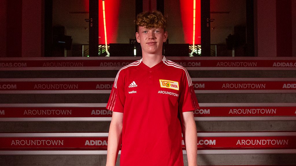 Junger Mann mit lockigem Haar trägt ein rotes Fußballtrikot von Union Berlin vor einer beleuchteten Kulisse.
