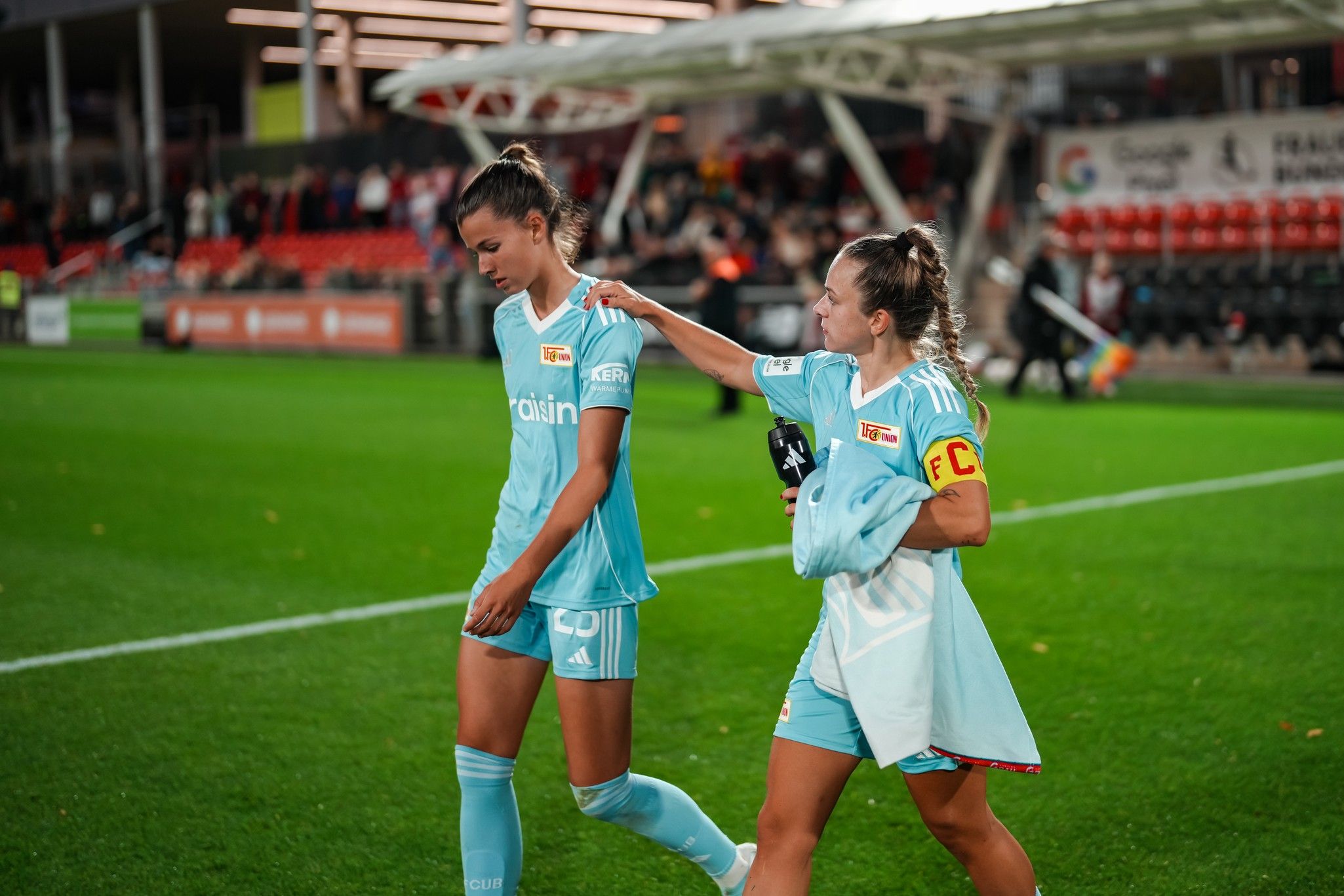 Zwei Fußballspielerinnen in blauen Trikots gehen auf dem Spielfeld, eine tröstet die andere. Zuschauer im Hintergrund.