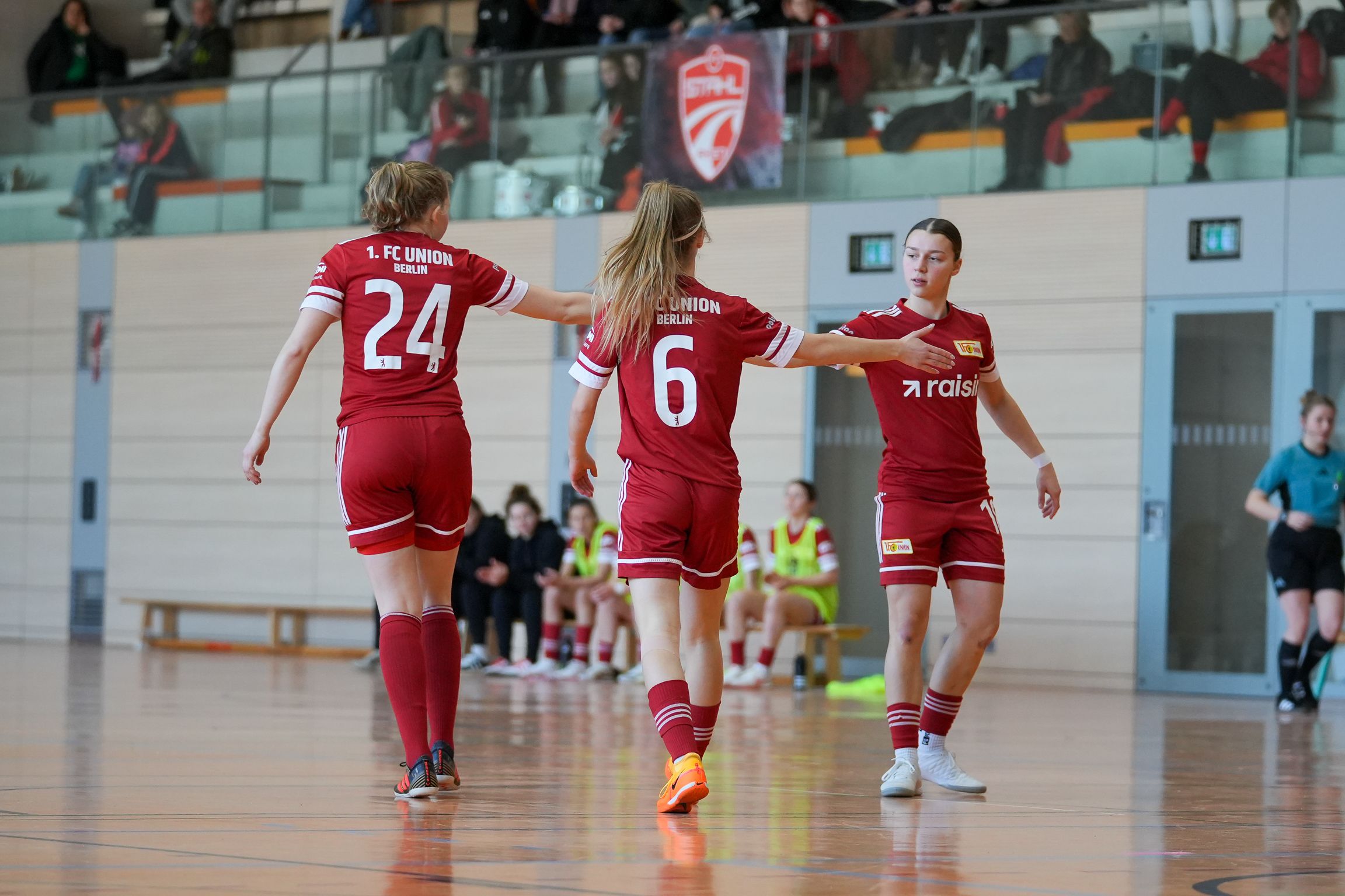 Drei Fußballspielerinnen in roten Trikots umarmen sich auf dem Spielfeld während eines Hallenspiels.