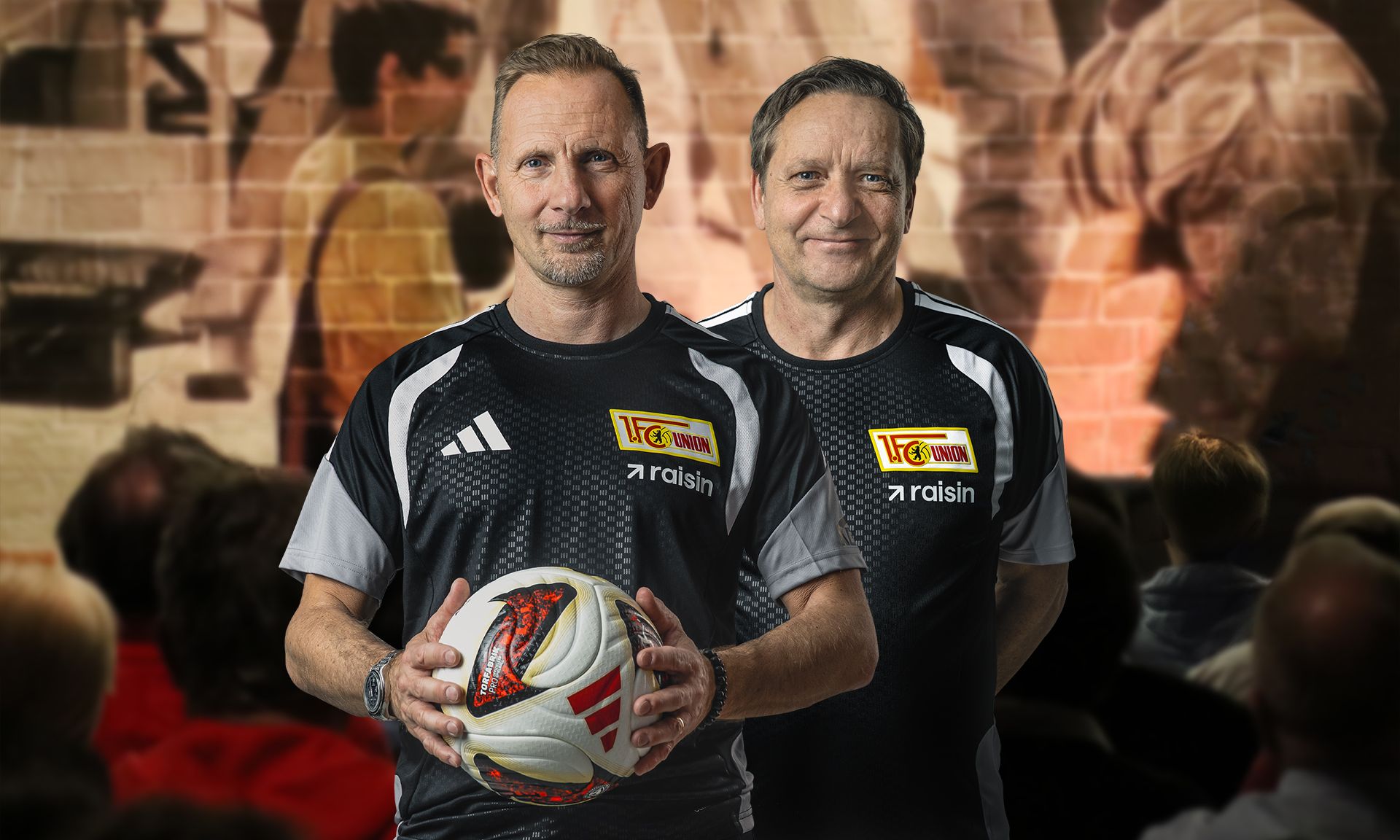 Zwei Männer in sportlichen Trikots halten einen Fußball, im Hintergrund ist eine unscharfe Menschenmenge zu sehen.