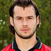 Porträt eines Mannes mit dunklen, langen Haaren in einem roten Sporttrikot, vor einer unscharfen grünen Hintergrundlandschaft.