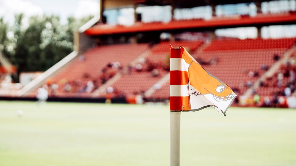 Ein Eckflagge mit einem orangefarbenen Motiv steht an einem leeren Fußballfeld vor einer Tribüne.