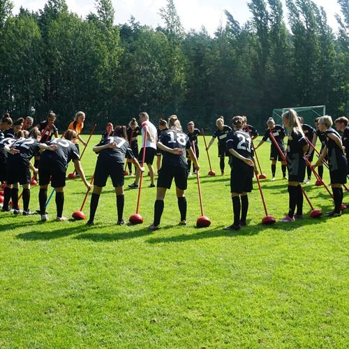 Eine Gruppe von Fußballspielerinnen steht auf einem Platz und hält Sportgeräte wie Besenstiele in der Hand.