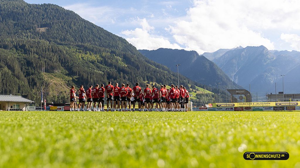 Fußballmannschaft trainiert auf einem grünen Platz mit Bergen im Hintergrund und blauem Himmel.