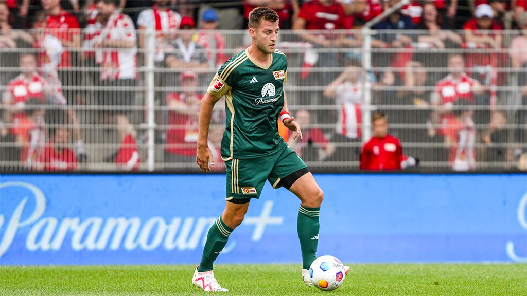 A soccer player in green goalkeeper gear stands on the field, with spectators in red jerseys in the background.
