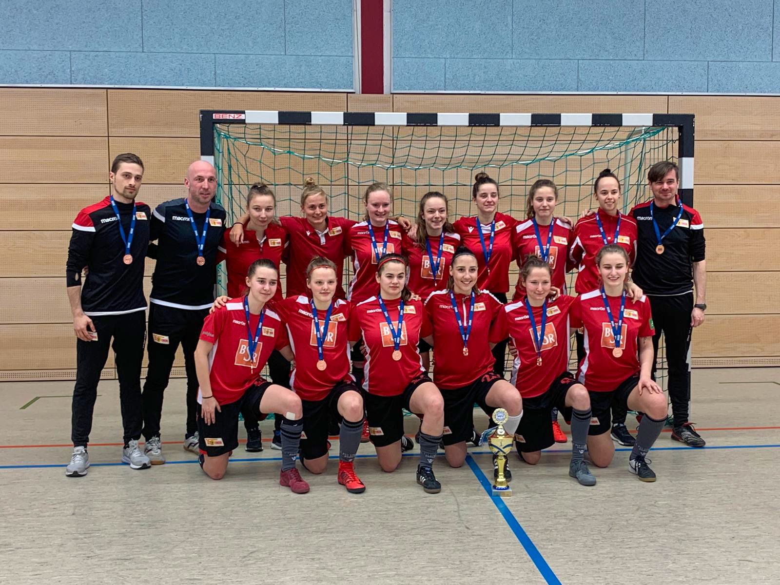 Eine Handballmannschaft in roten Trikots posiert mit Medaillen und einem Pokal vor einem Tor.