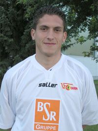 Ein Mann mit kurzen Haaren trägt ein weißes Fußballtrikot mit Logos und steht vor einem Baum.