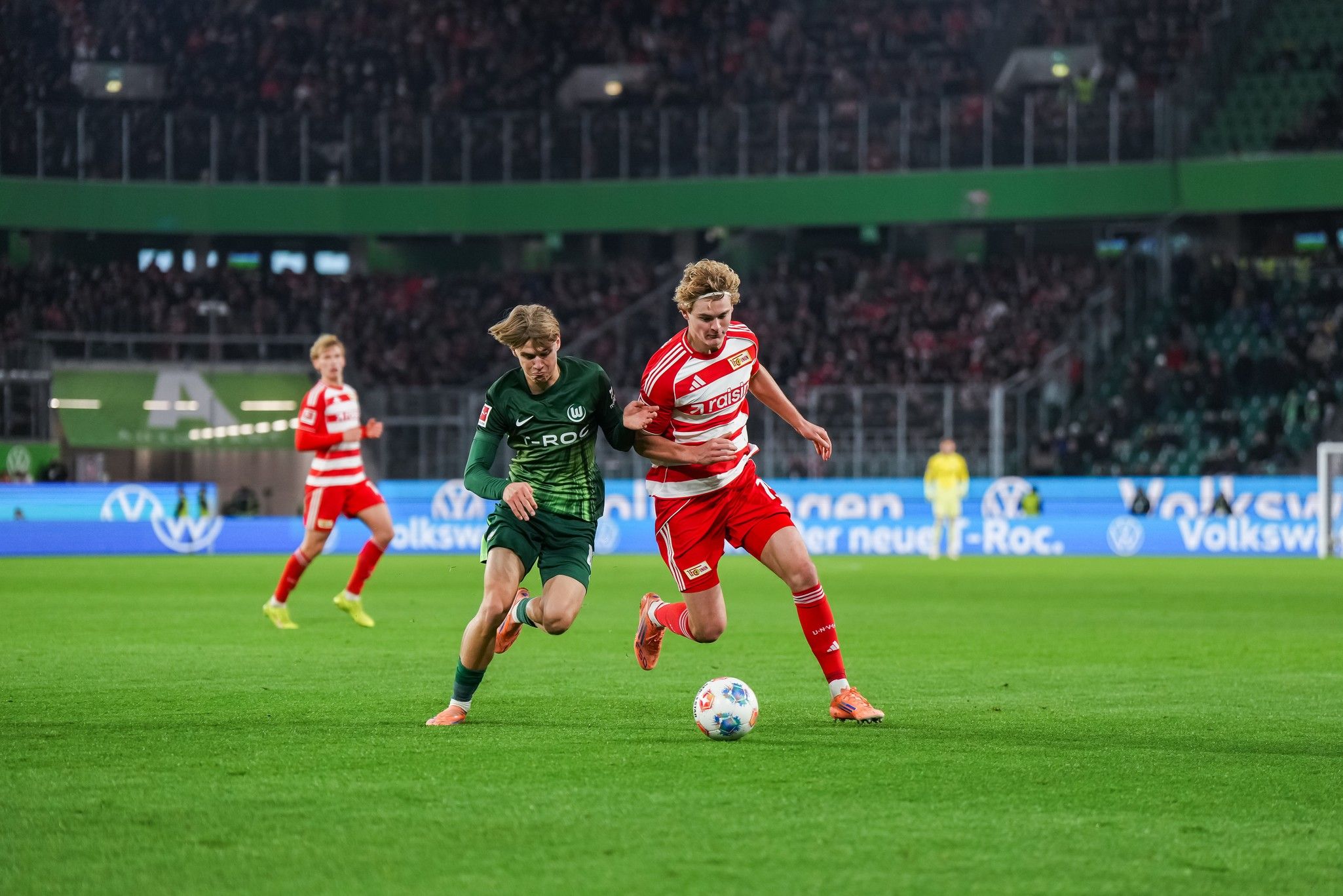 Zwei Fußballspieler im Wettkampf um den Ball während eines Spiels in einem Stadion mit jubelndem Publikum.