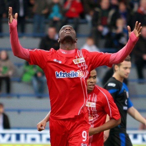 Ein Fußballspieler in rotem Trikot hebt die Hände zum Jubel, während ein weiterer Spieler im Hintergrund steht.