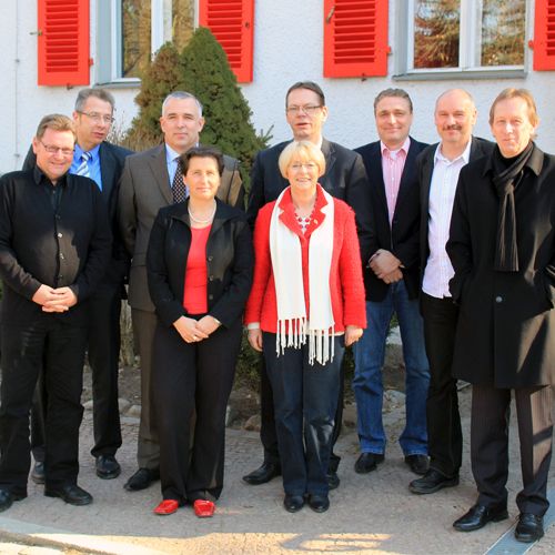 Eine Gruppe von neun Personen steht vor einem Gebäude mit roten Fensterläden. Sie lächeln und posieren gemeinsam für das Foto.