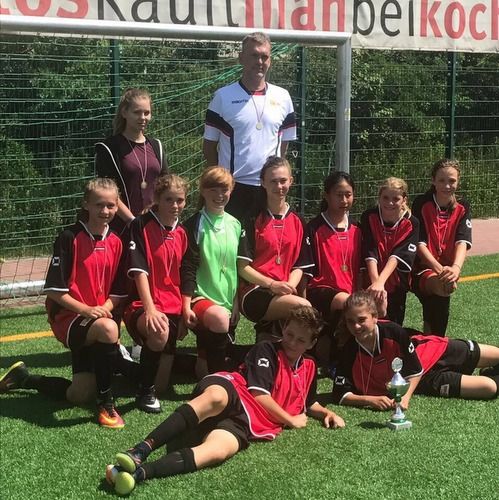 Eine Mädchenfußballmannschaft in roten Trikots posiert mit einem Pokal und ihrem Trainer auf einem Kunstrasenplatz.