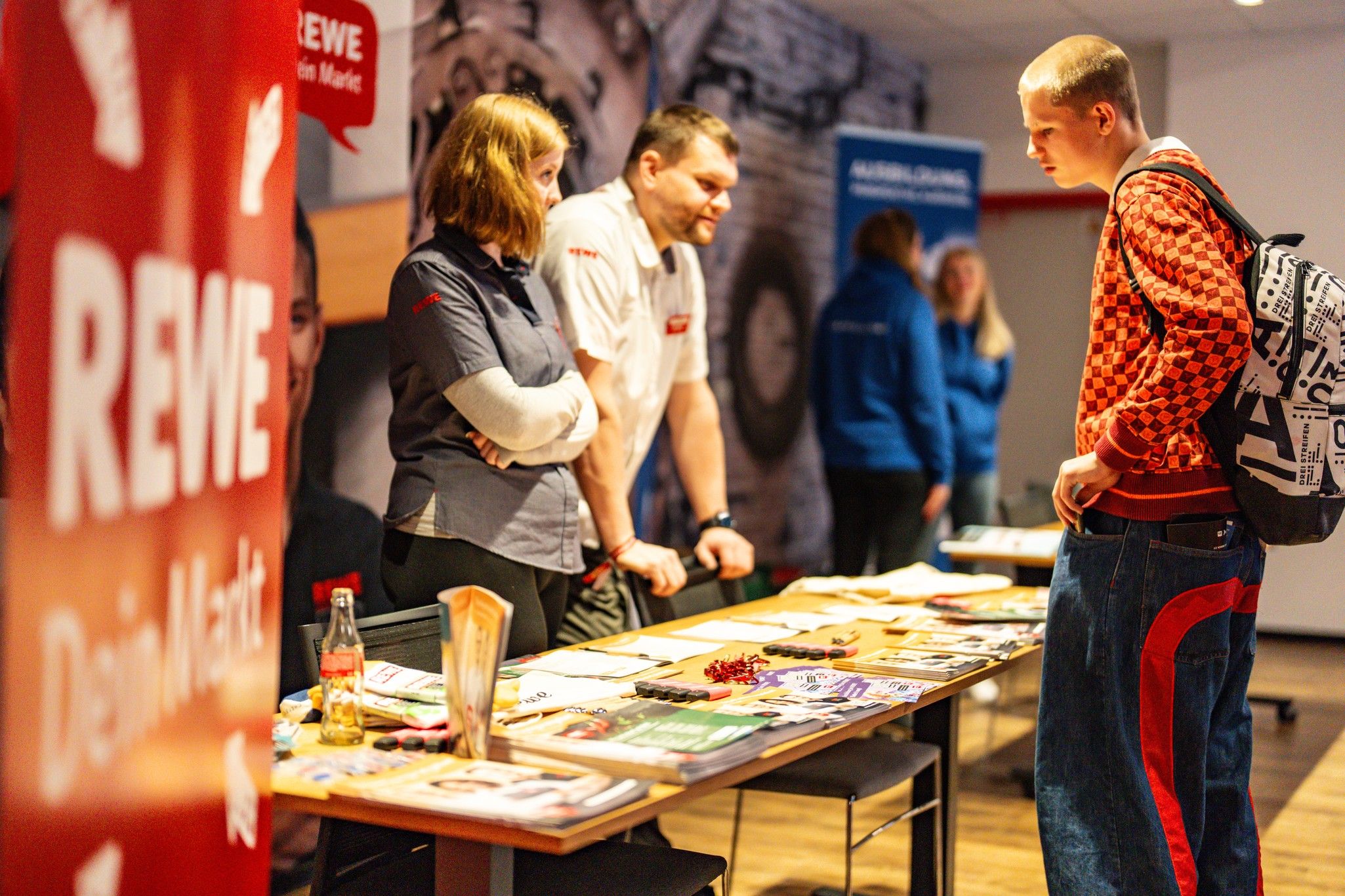 Messe mit einem Stand von REWE, zwei Personen im Gespräch und ein junger Besucher, der sich Informationsmaterial ansieht.