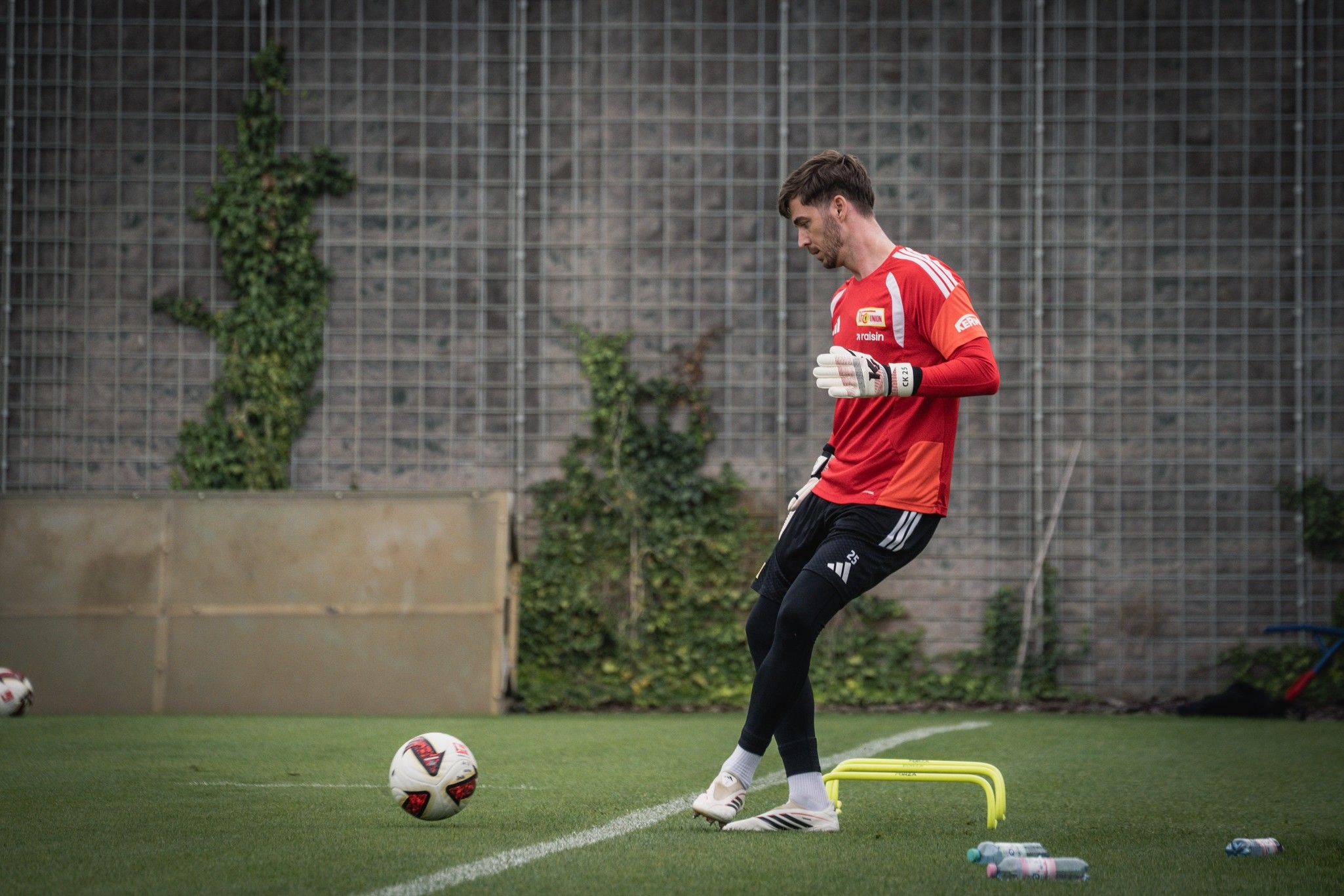 Torwart in rotem Trainingsshirt übt auf einem Fußballplatz, mit Hütchen und Wasserflaschen im Hintergrund.