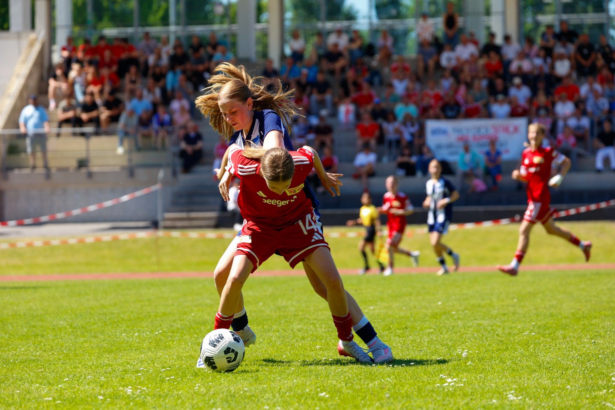 Zwei Kinder spielen Fußball auf einem Feld; ein Spieler wird von einem anderen gefasst. Zuschauer sind im Hintergrund sichtbar.