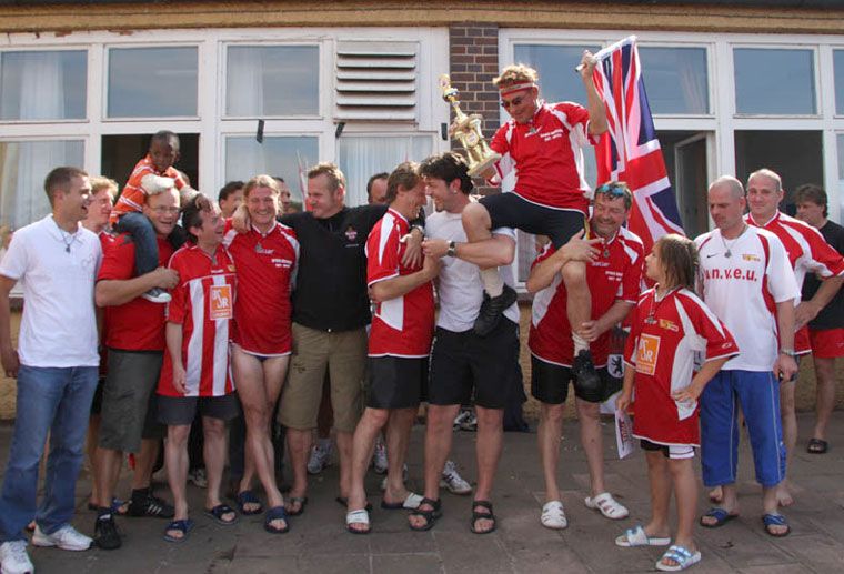 Menschen in roten T-Shirts feiern mit einer Trophäe und einer britischen Flagge, umgeben von einem Haus und anderen Teilnehmern.