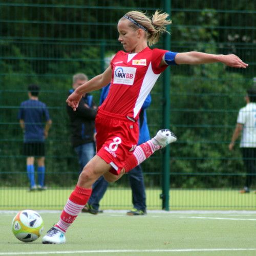 Eine Spielerin in rotem Fußballtrikot schießt den Ball auf einem Sportplatz, im Hintergrund sind Zuschauer zu sehen.