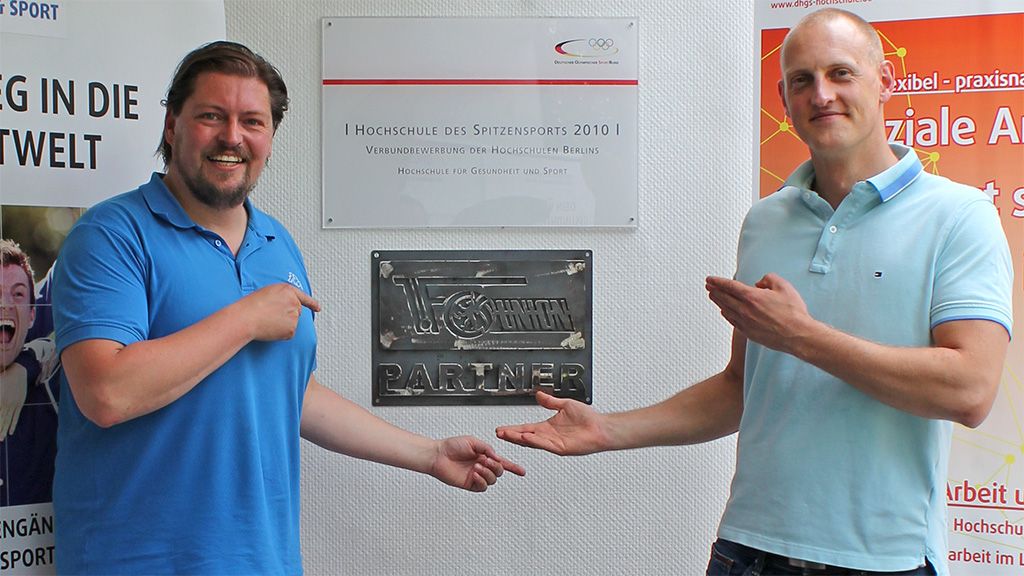 Zwei Männer zeigen auf ein Schild mit der Aufschrift "Partner" in einem Raum der Hochschule für Gesundheit und Sport.