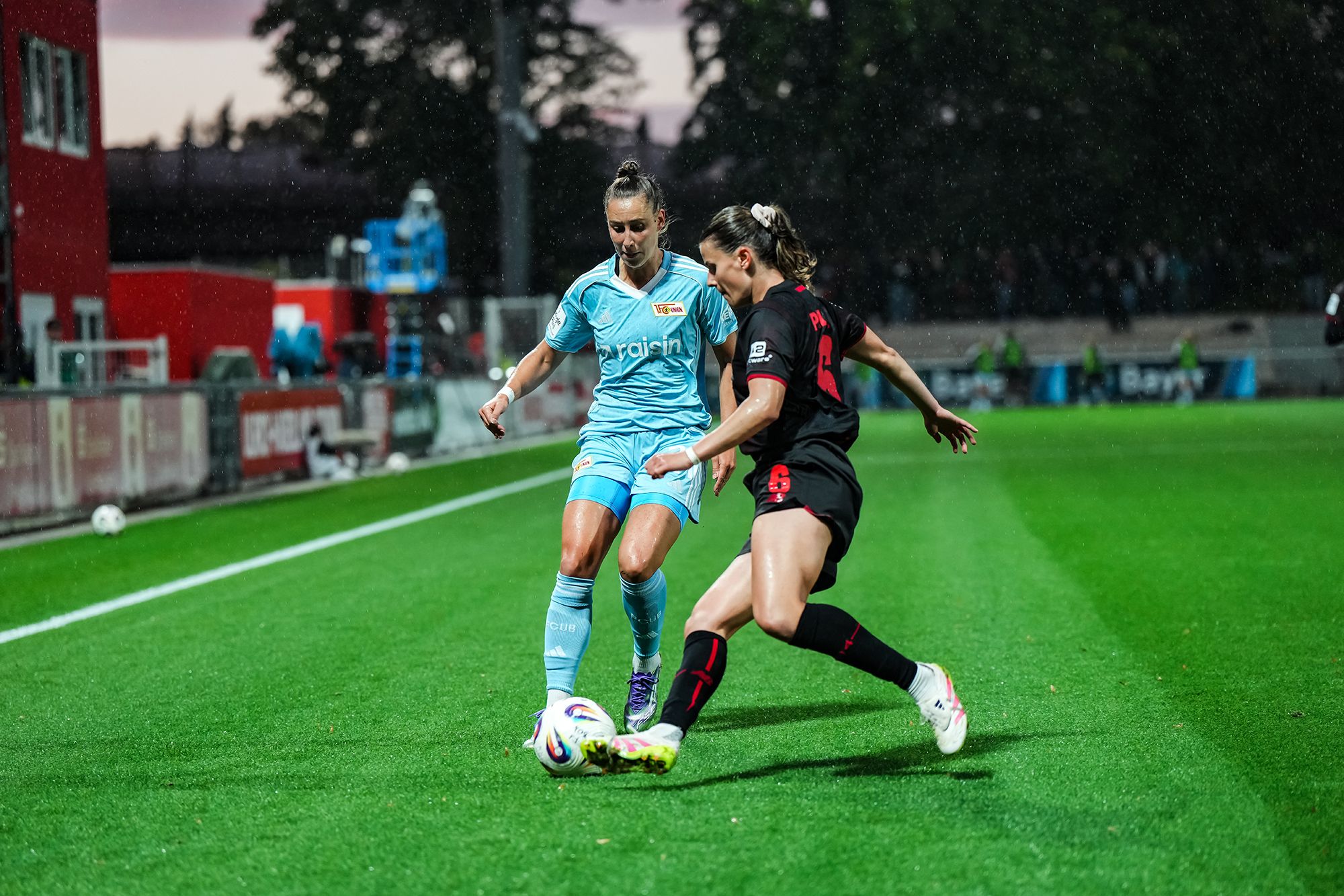 Zwei Fußballspielerinnen im Wettkampf um den Ball auf einem Rasenplatz bei Abenddämmerung.
