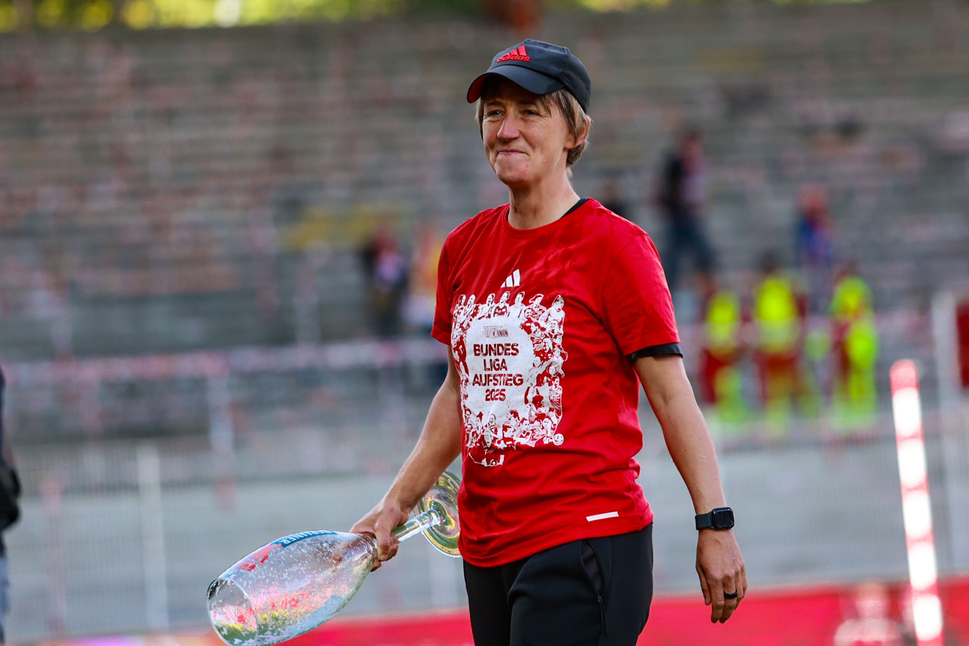 Eine Person in rotem Shirt und schwarzem Cap hält eine große Flasche und lächelt, während sie auf einem Sportfeld steht.